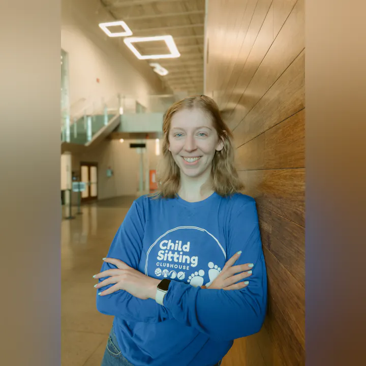 A smiling individual in a blue shirt stands against a wooden wall, promoting "Child Sitting Clubhouse" in a modern space.