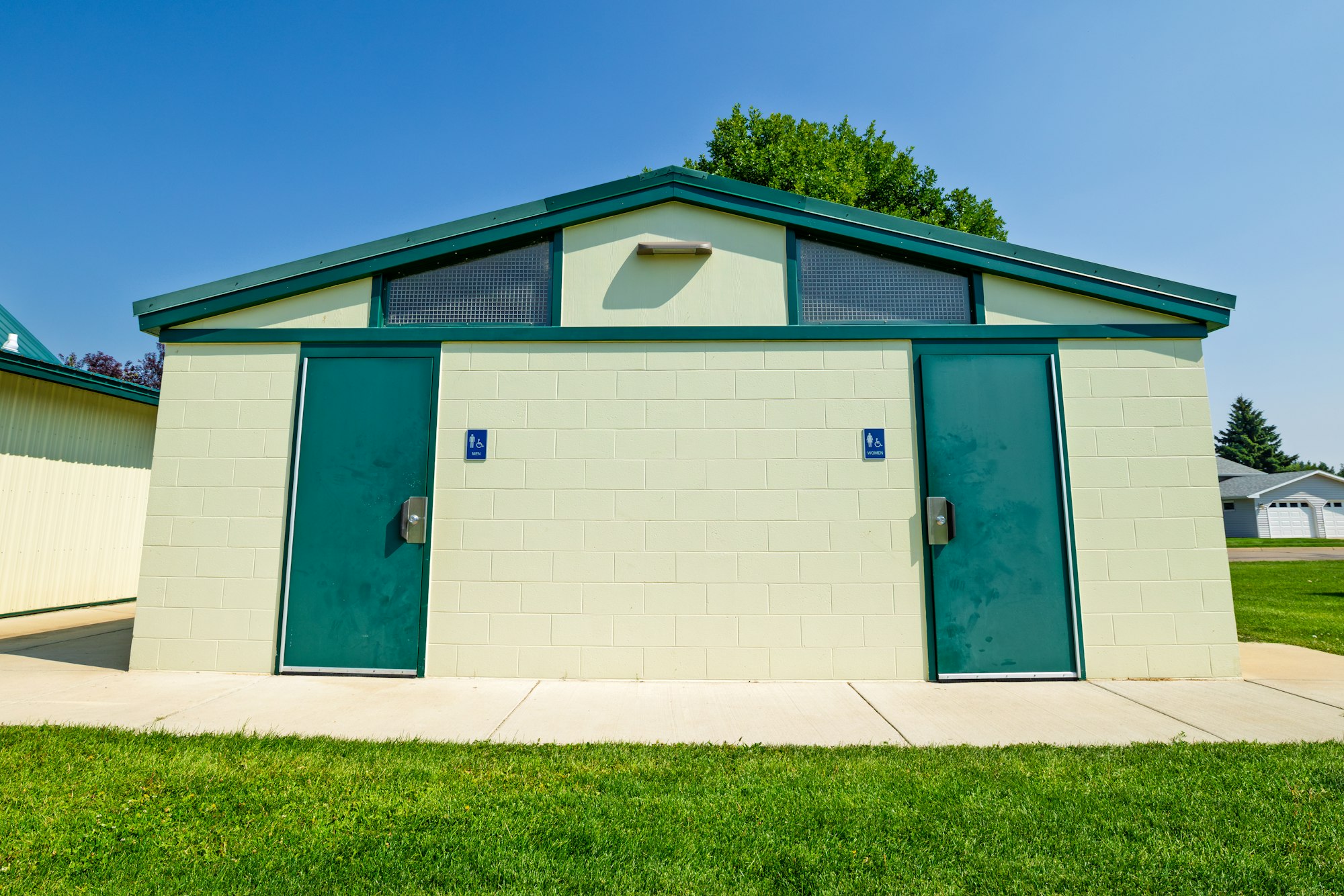 The image shows a public restroom building with two separate entrances, set against a clear blue sky and green grass.