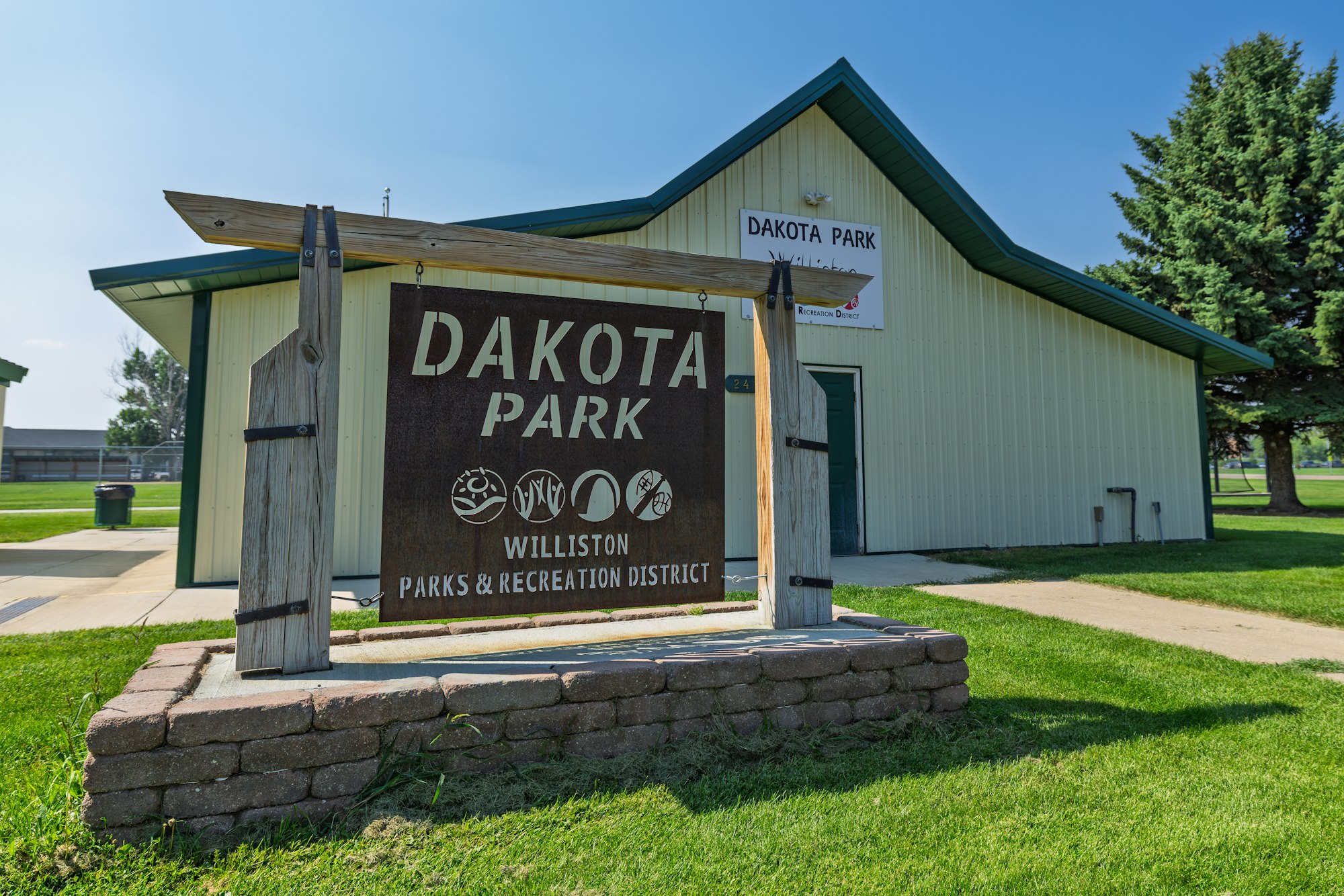 The image shows a sign for Dakota Park in Williston, featuring a building and grassy area. It's part of the local Parks & Recreation District.