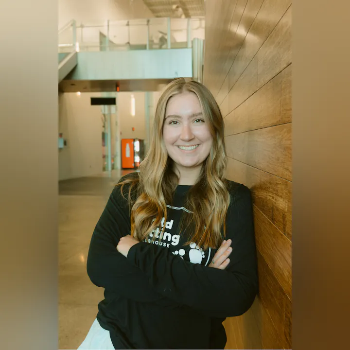 A smiling individual with long hair leans against a wooden wall, wearing a black shirt with a logo on it.