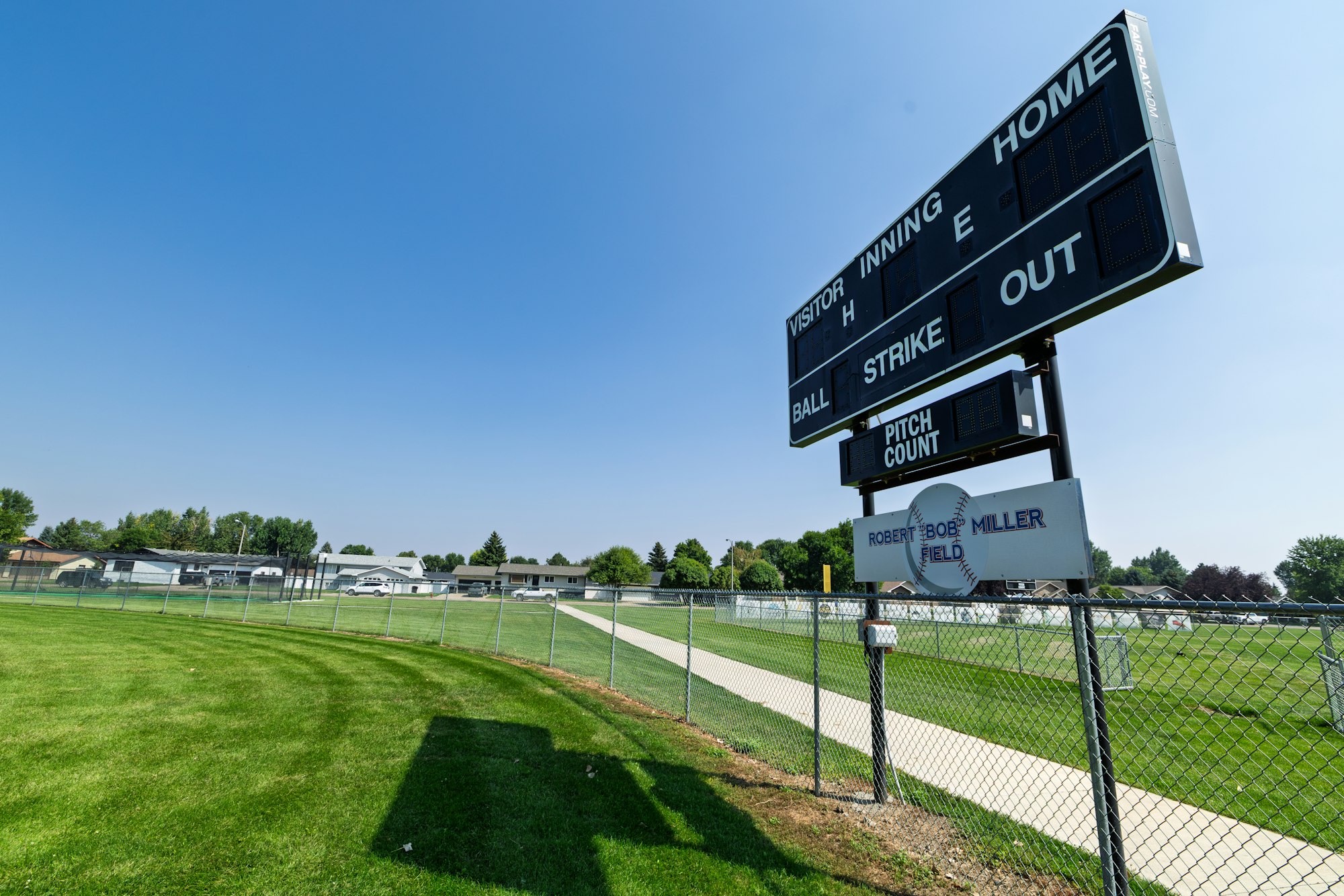 A baseball field scoreboard shows game stats, including innings, strikes, and balls, set against a clear blue sky.