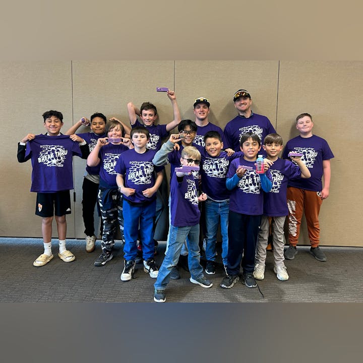 A group of boys and two adults wearing matching purple "Break Thru" shirts, smiling and holding items, posing indoors.