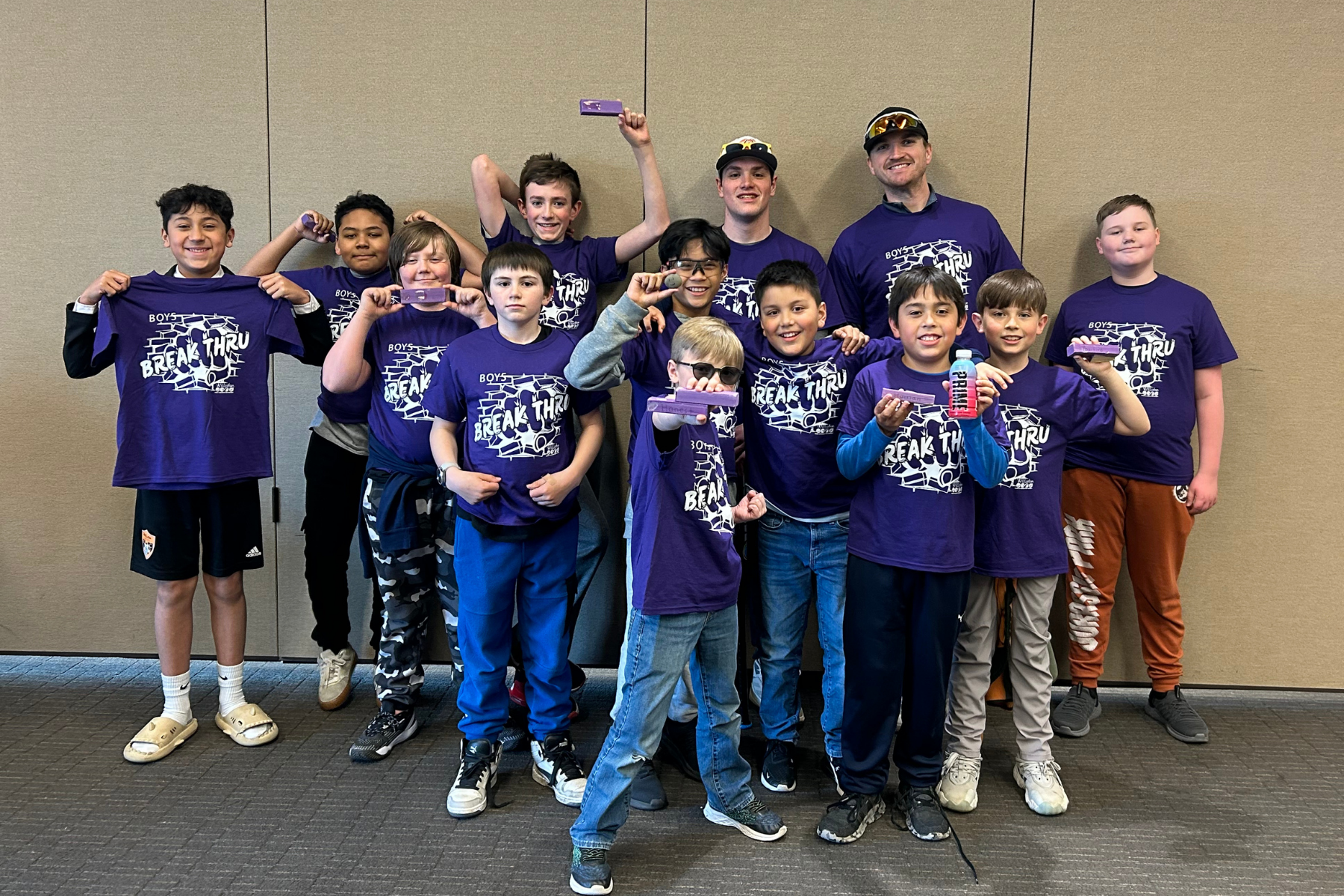 A group of boys and two adults wearing matching purple "Break Thru" shirts, smiling and holding items, posing indoors.