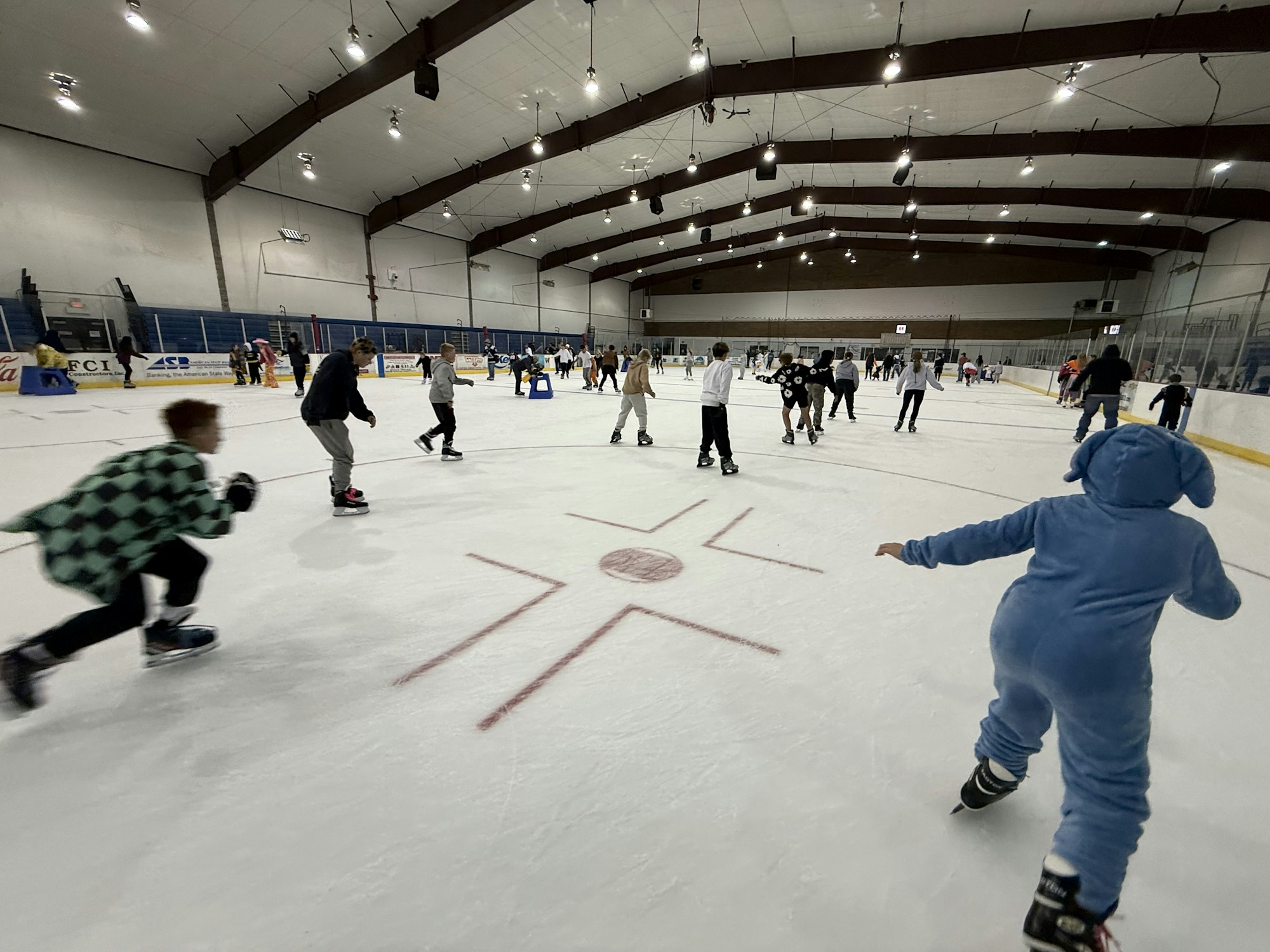 A bustling ice rink filled with people skating, including one in a blue onesie, under a spacious arena ceiling.