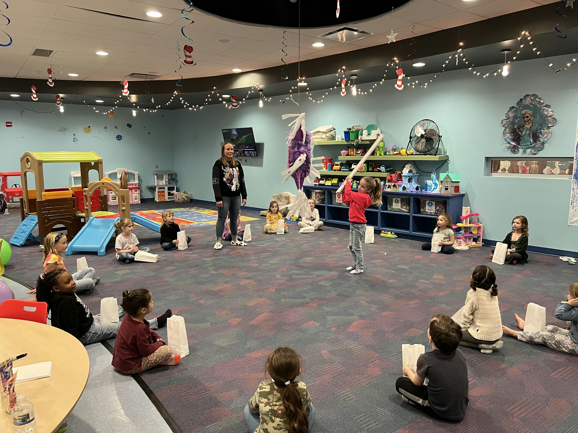 Children in a playroom with a piñata and an adult supervising. Festive decorations are visible.
