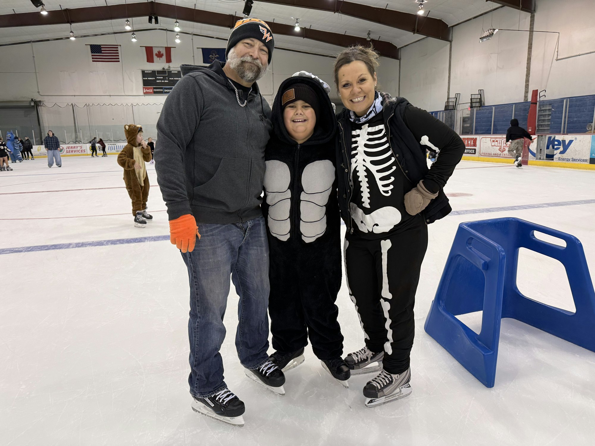 A family enjoys ice skating, dressed in fun costumes: a skeleton and a bear, with a background of skaters and flags.