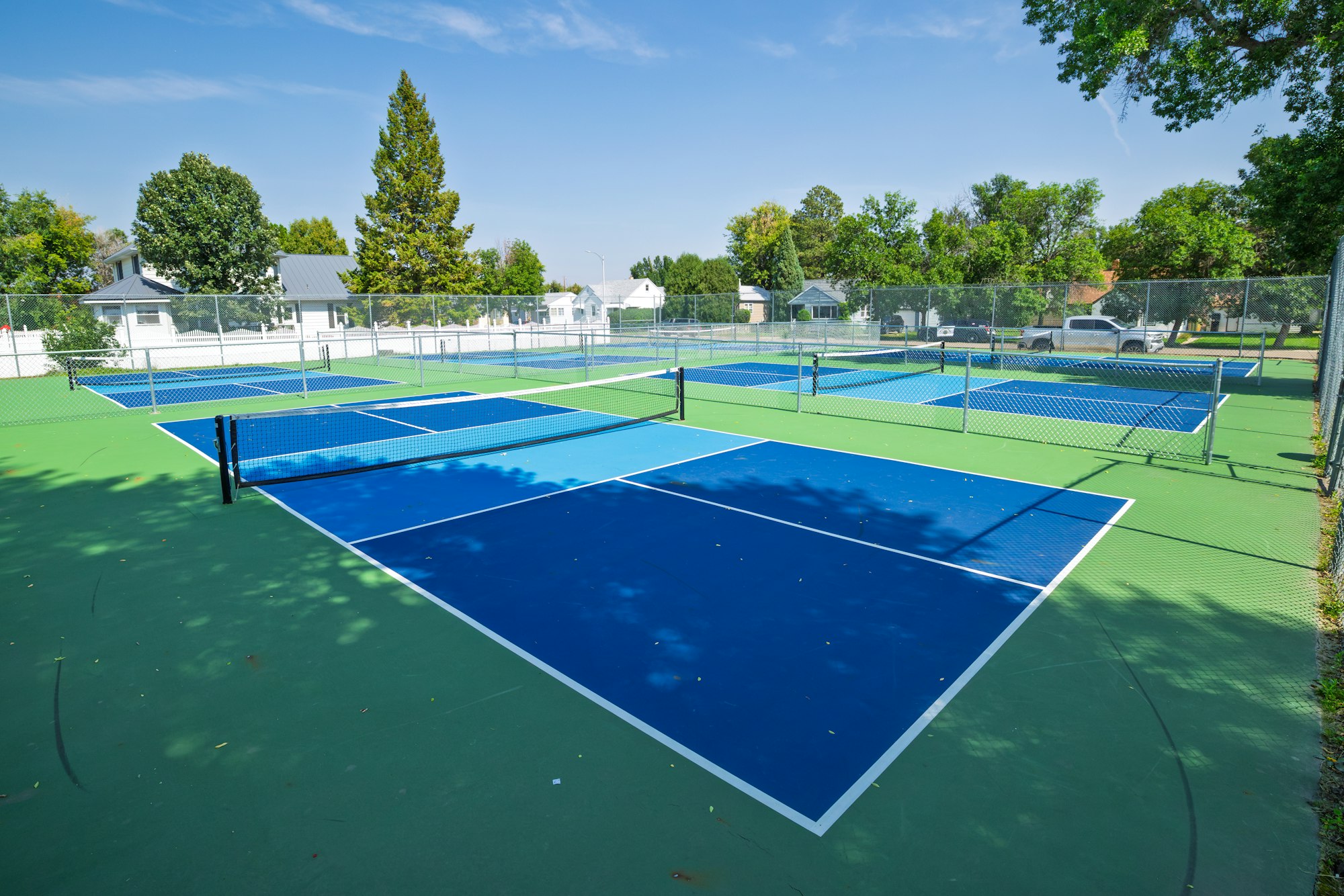 The image shows a bright, sunny tennis court area with multiple blue courts and surrounding greenery and homes.