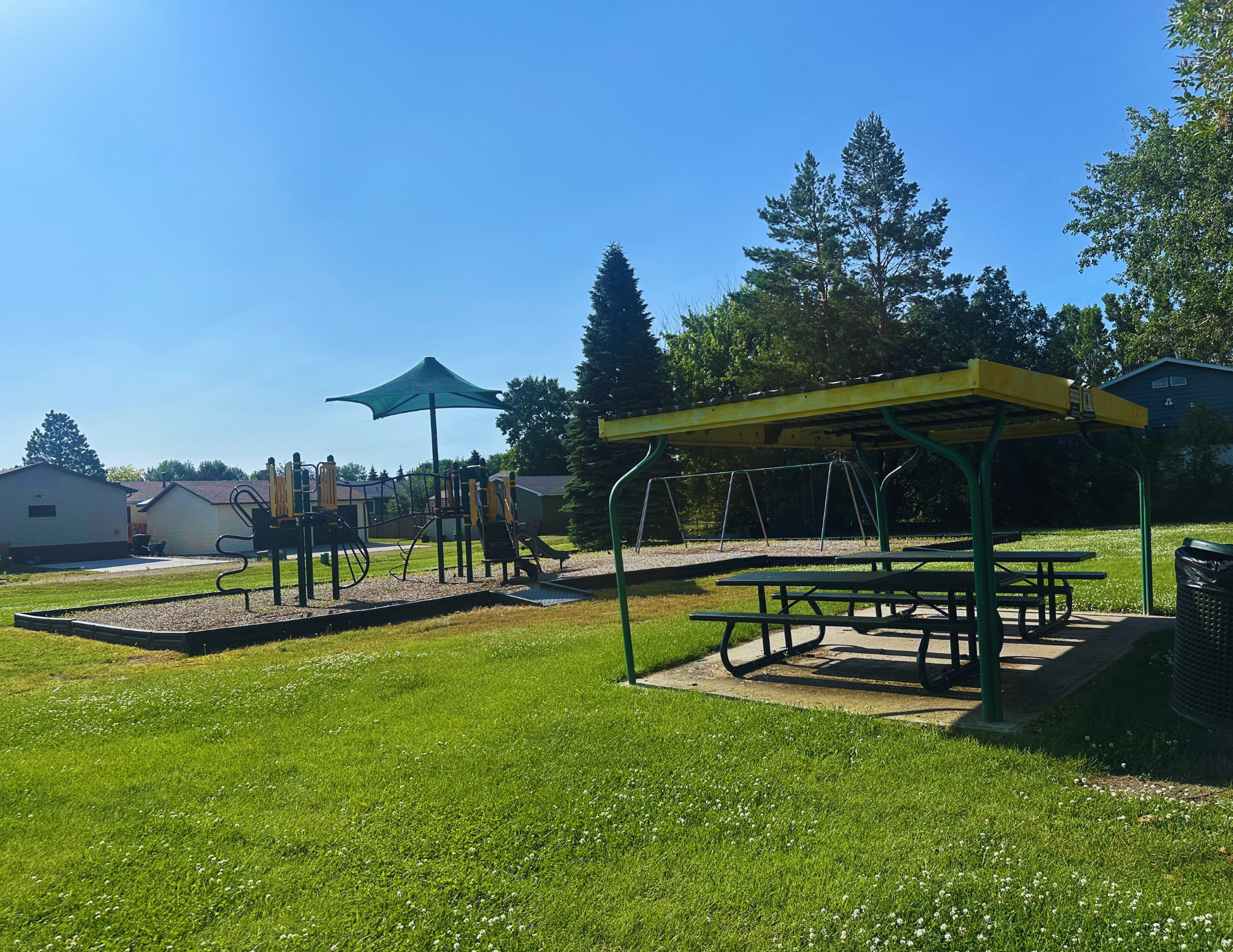 Playground with slides and swings, covered picnic tables, green grass, trees, and houses in the background under a clear blue sky.