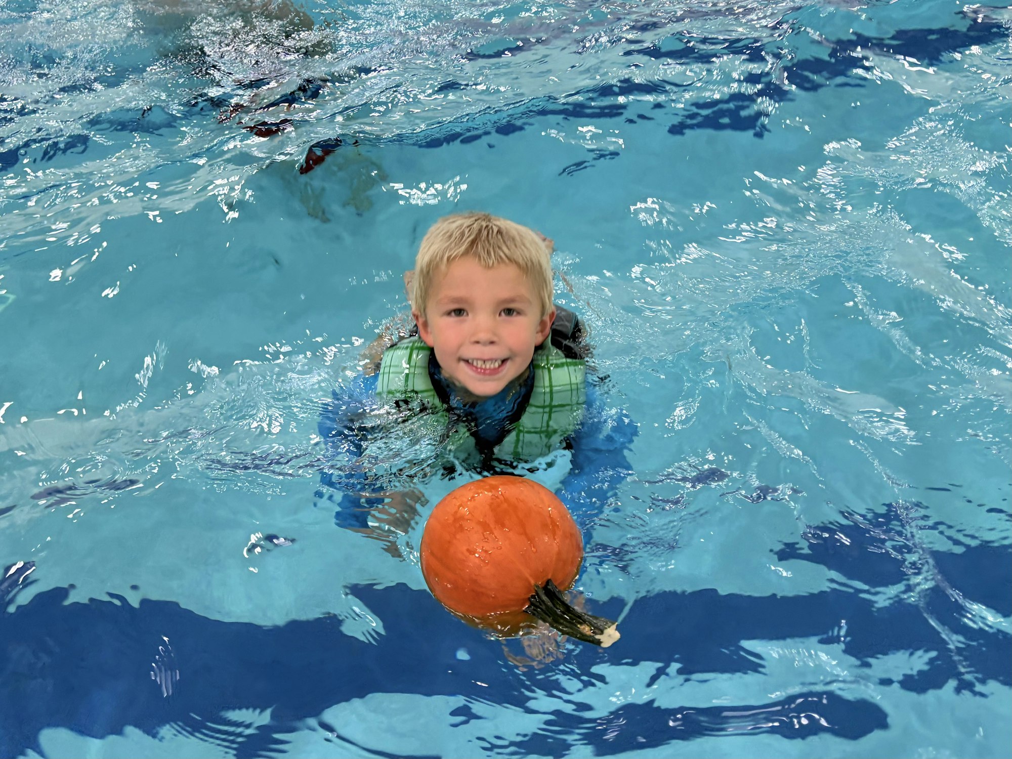 A smiling child in a swimming pool holds a pumpkin in the water while wearing a life jacket.