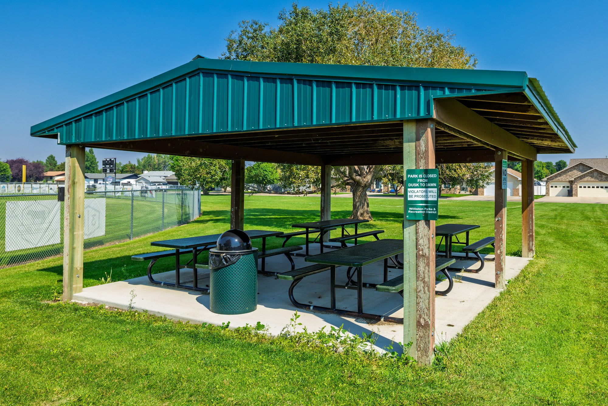 A shaded picnic area with metal tables and a trash can, surrounded by grass and a fence, with a park sign nearby.