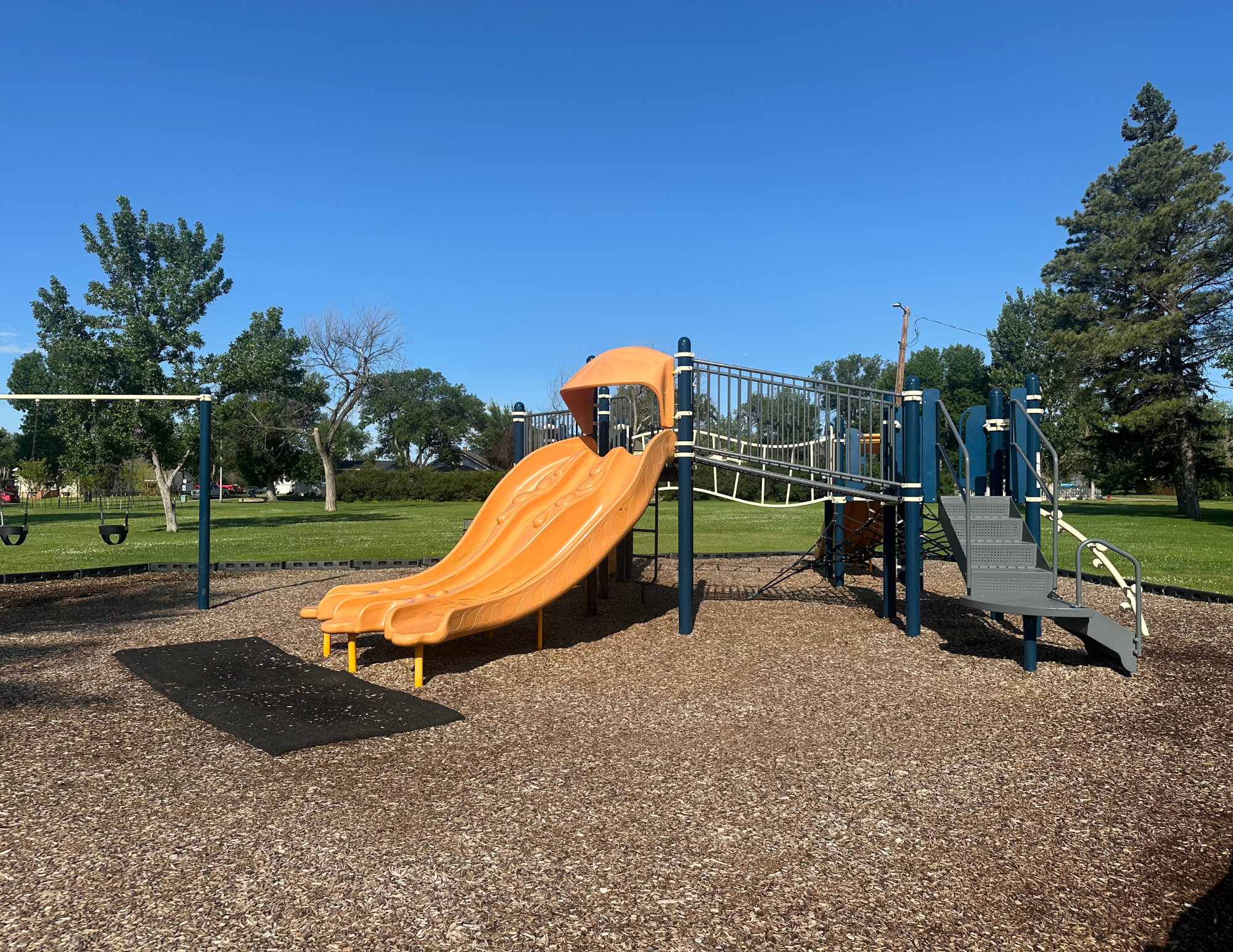 Playground with slides, swings, climbing equipment, trees, and green grass under a clear blue sky.