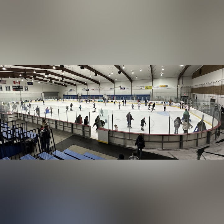 An indoor ice skating rink with people skating and spectators watching from the sidelines.