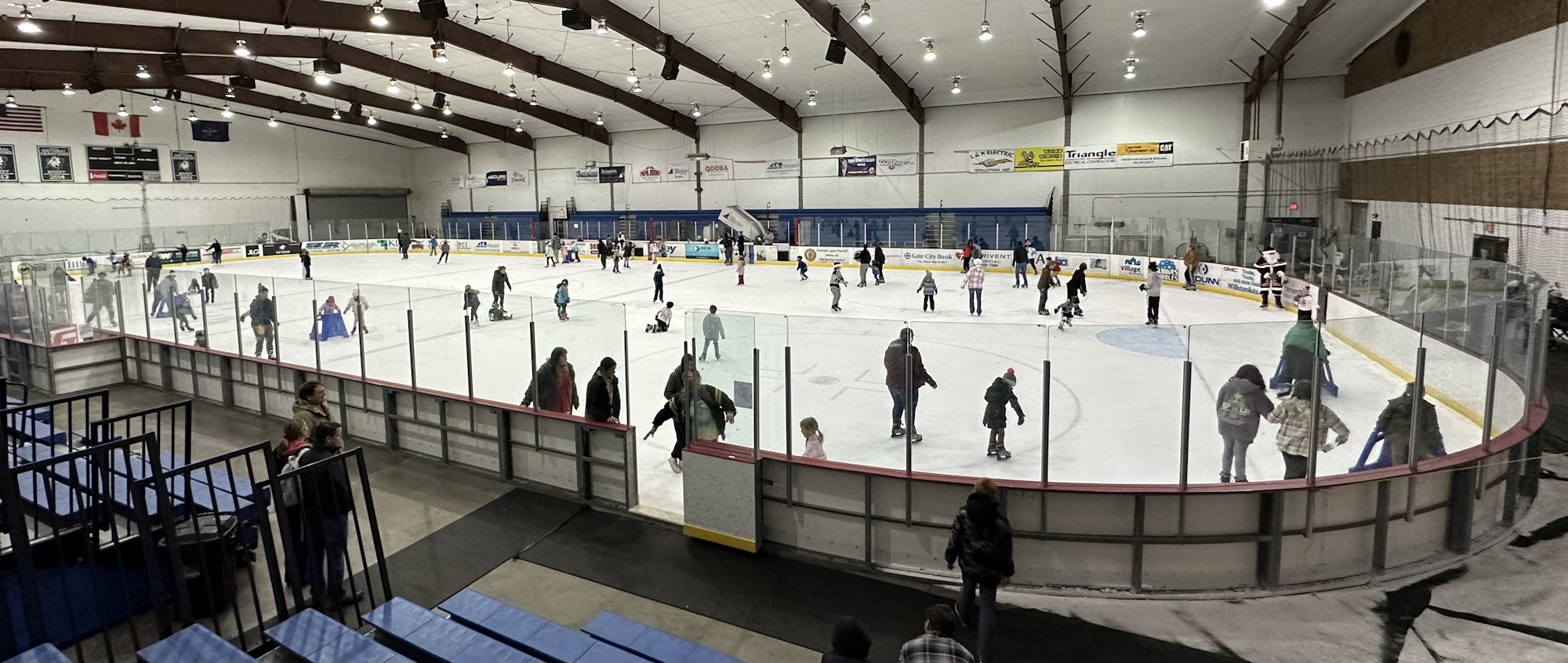 An indoor ice skating rink with people skating and spectators watching from the sidelines.