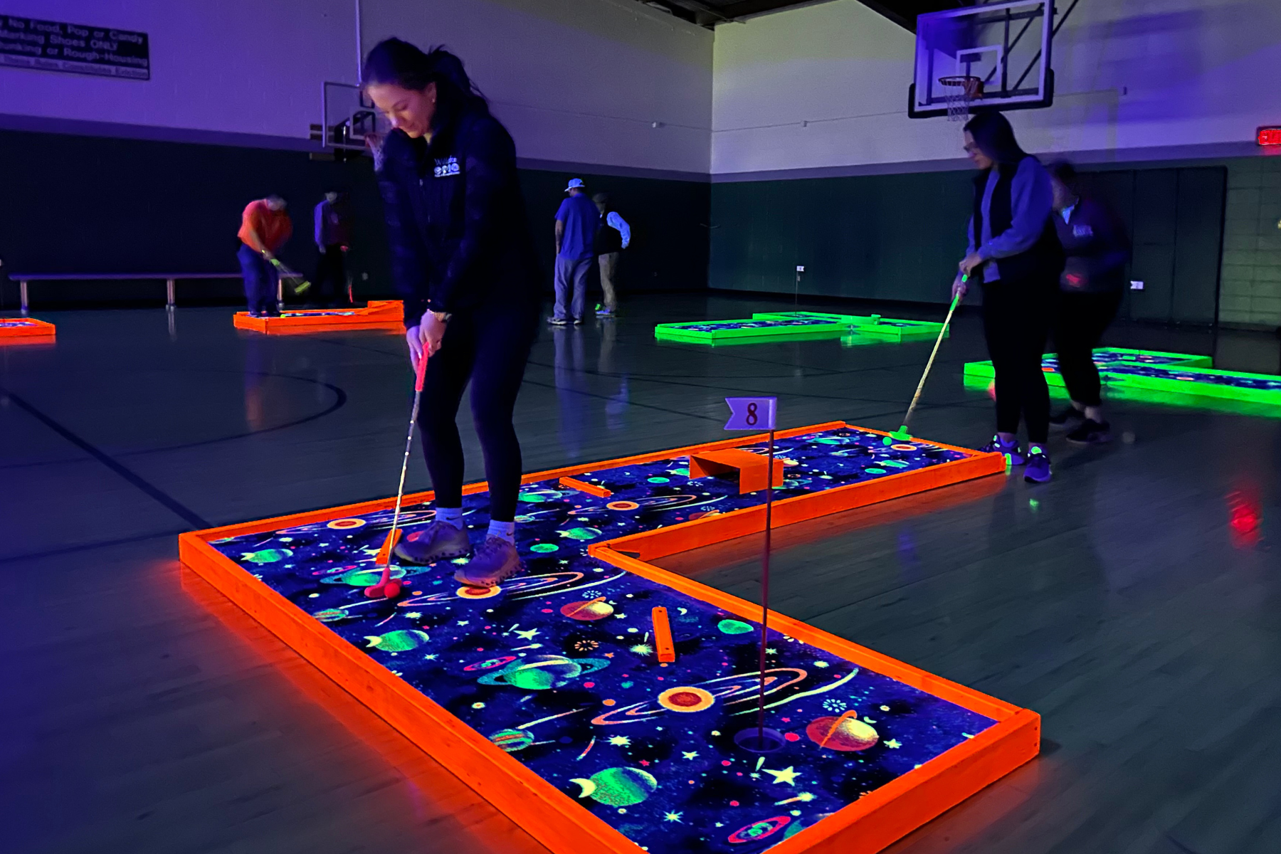People playing glow-in-the-dark mini golf in a dark room with space-themed designs on the courses.