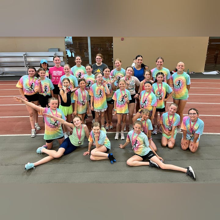 A group of girls and coaches in colorful tie-dye shirts pose indoors, some with medals, on a track.