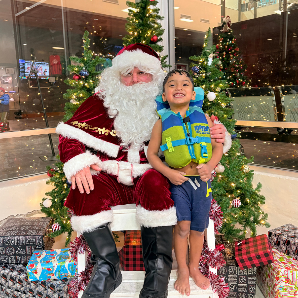 A joyful child in a life jacket sits on Santa's lap, surrounded by Christmas trees and festive decorations.
