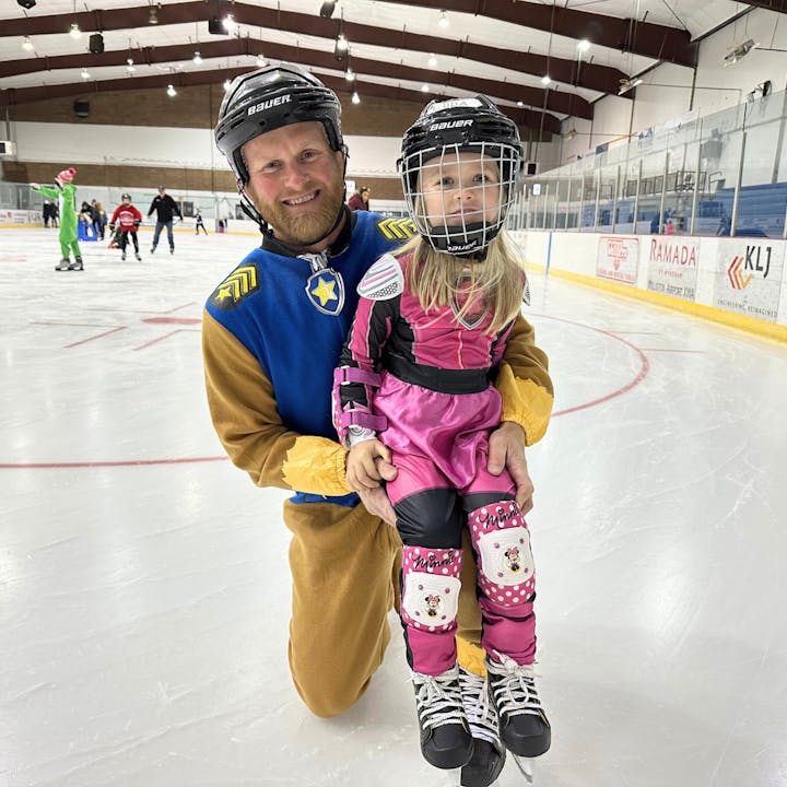 Adult and child on ice rink with skates and helmets, smiling.