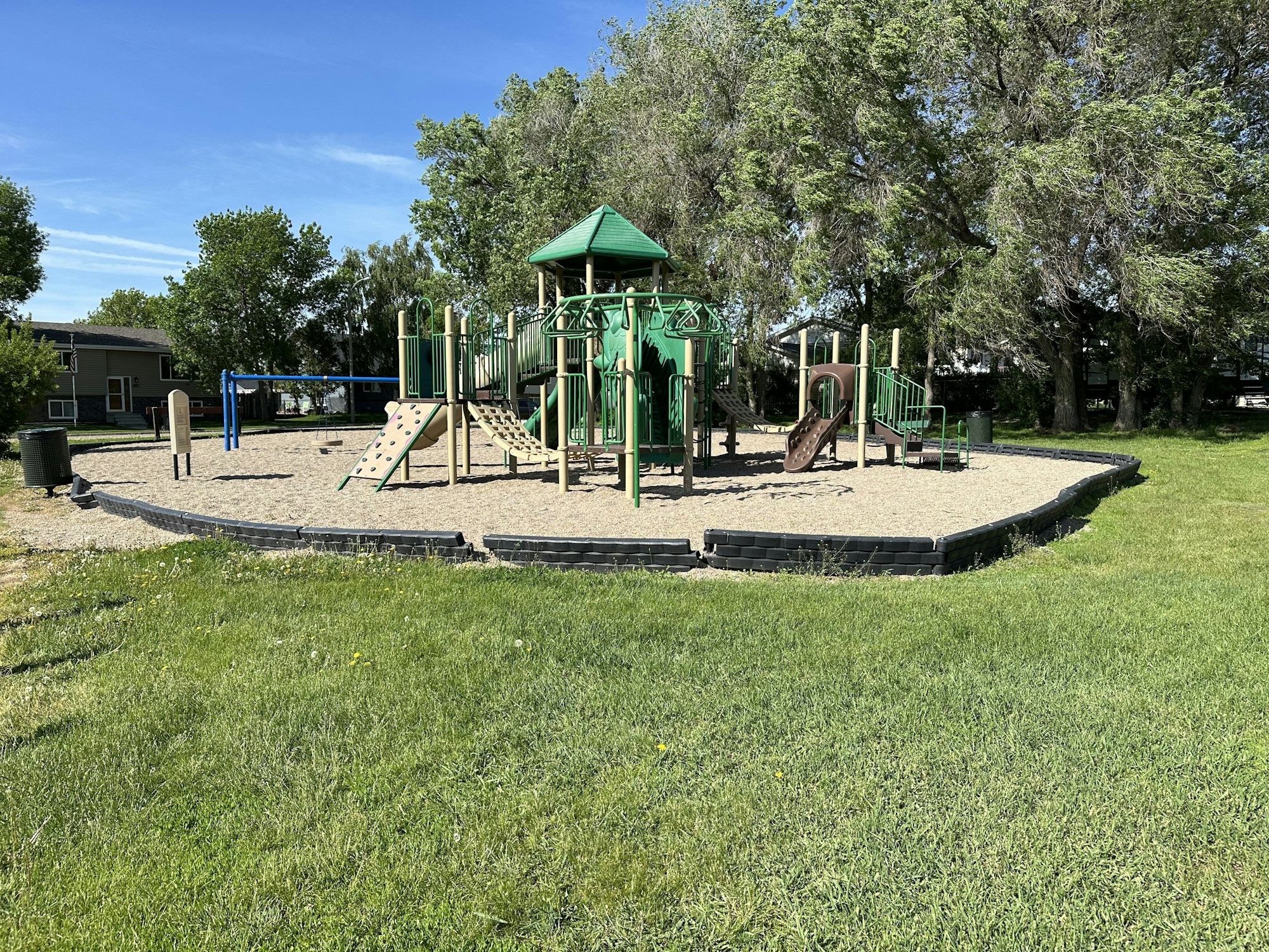 A playground with swings and climbing equipment encircled by sand and grass, surrounded by trees.