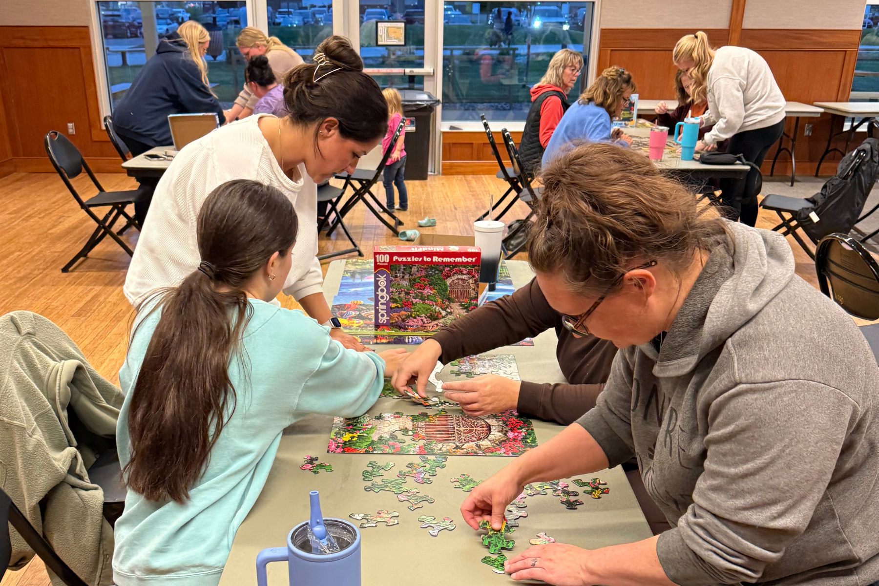 People are working together on a jigsaw puzzle in a community space, with multiple tables and engaging activity.