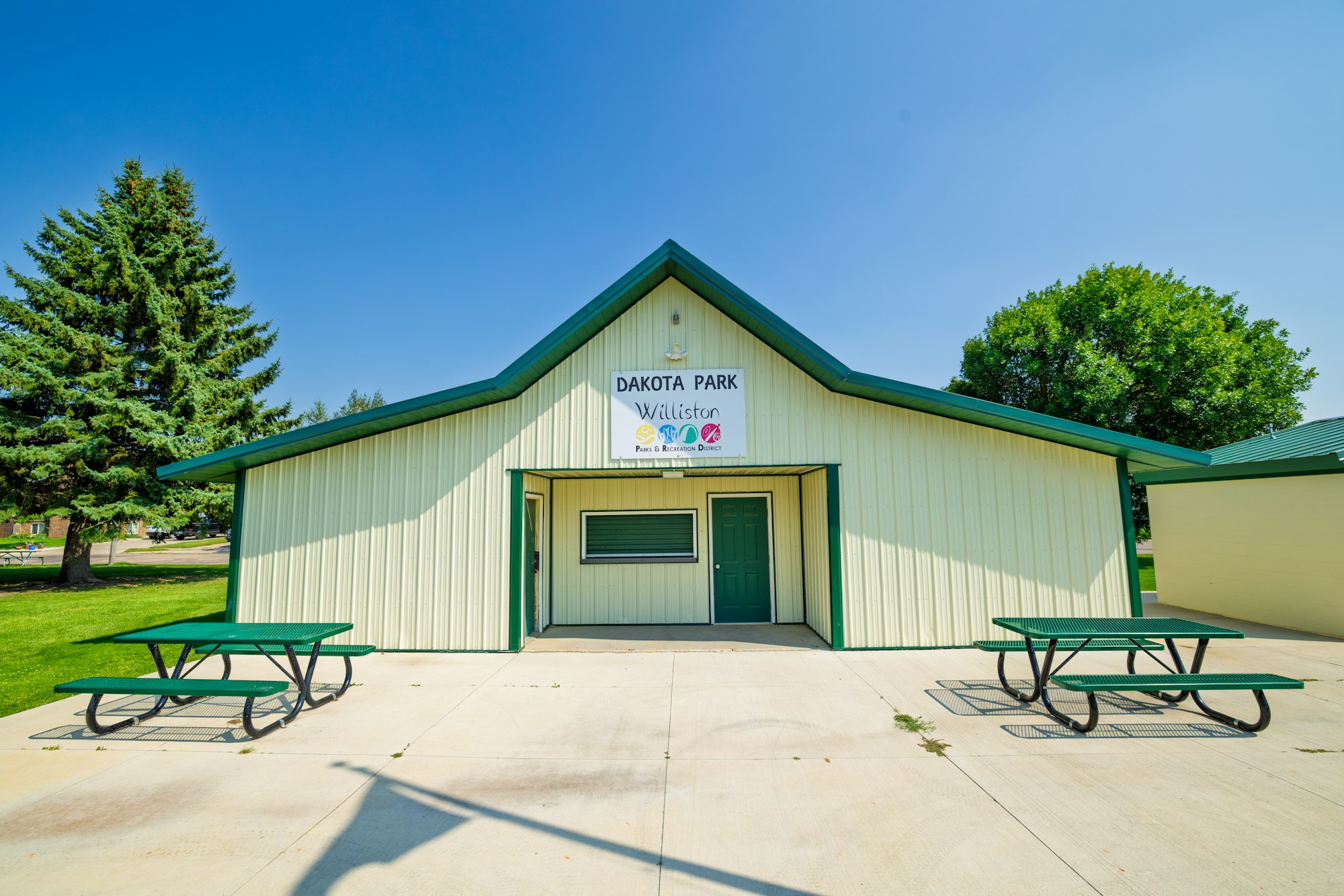 A community building at Dakota Park in Williston, featuring picnic tables and green landscaping under a clear blue sky.