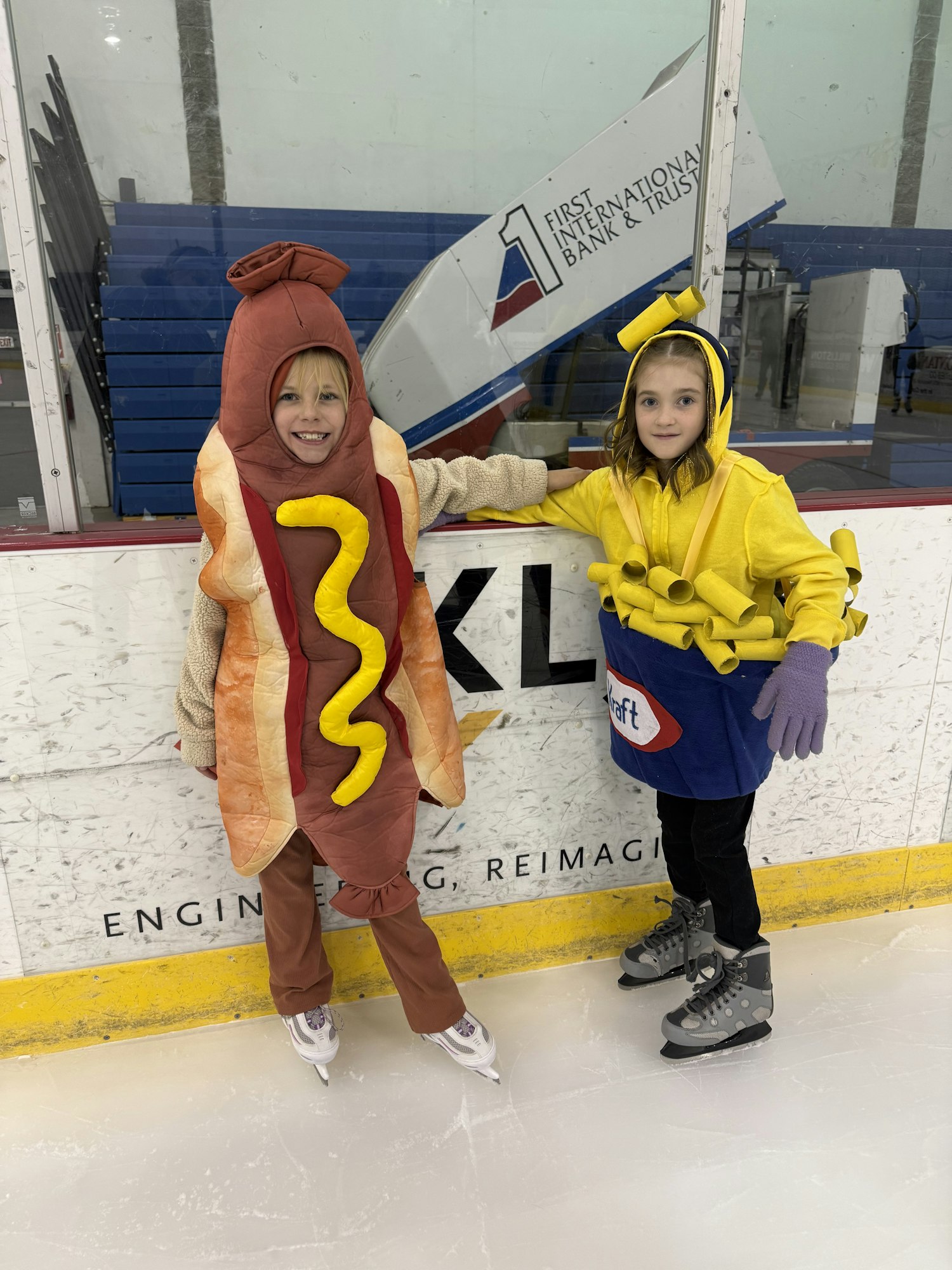 Two kids in playful food costumes (a hot dog and a banana) are on an ice rink, ready for fun.