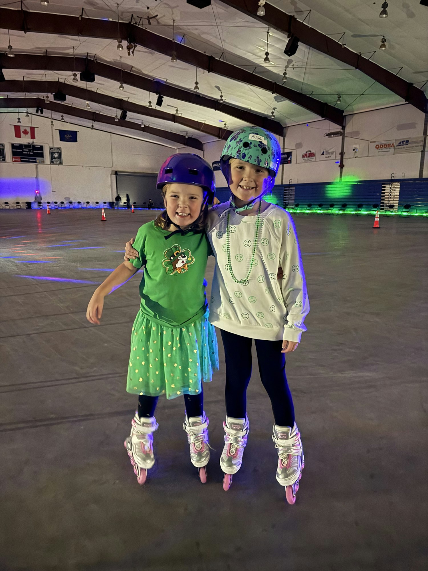 Two kids in green outfits and helmets are roller skating in an indoor rink.