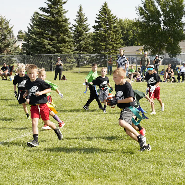 Kids playing flag football in a grassy field, with spectators and trees in the background.