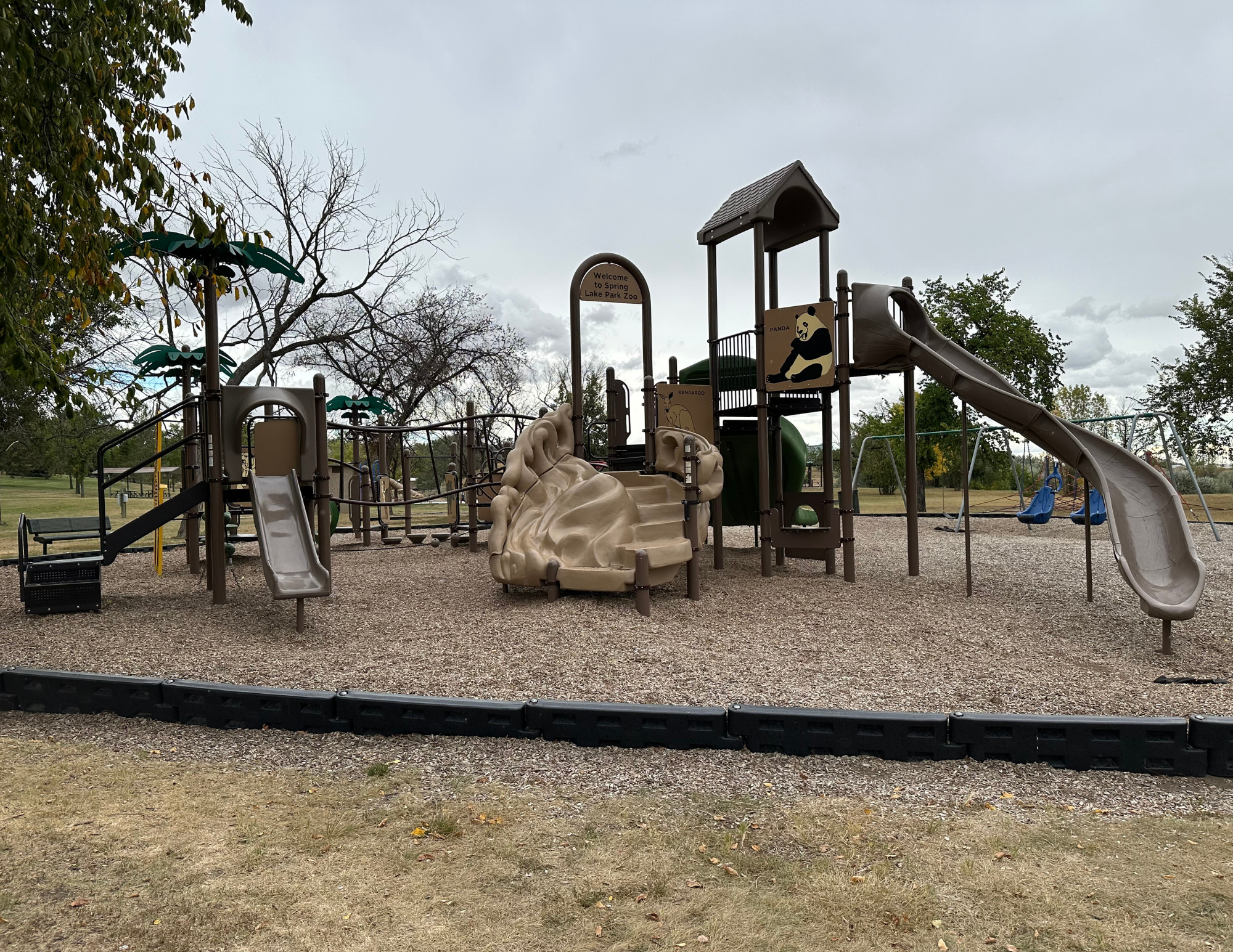A playground with slides, climbing elements, swings, and play structures, set in a park with trees.