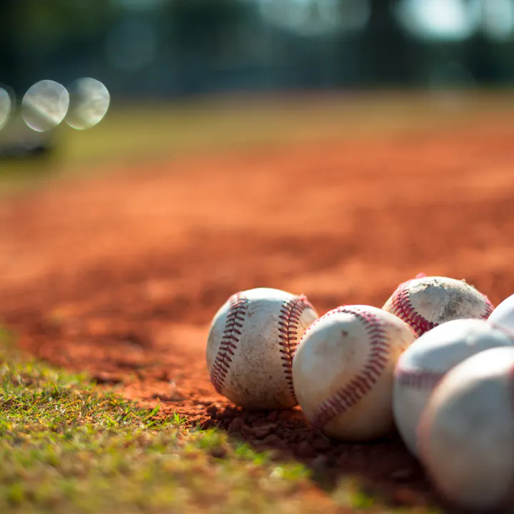 A close-up view of several dirty baseballs resting on a sandy field, with a blurred background and sunlight.