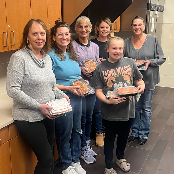 A group of six women and a girl are smiling while holding food containers in a kitchen setting.