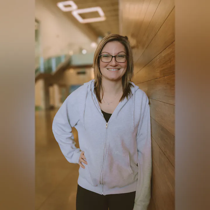 A smiling person wearing glasses and a gray hoodie stands against a wooden wall in a well-lit space.