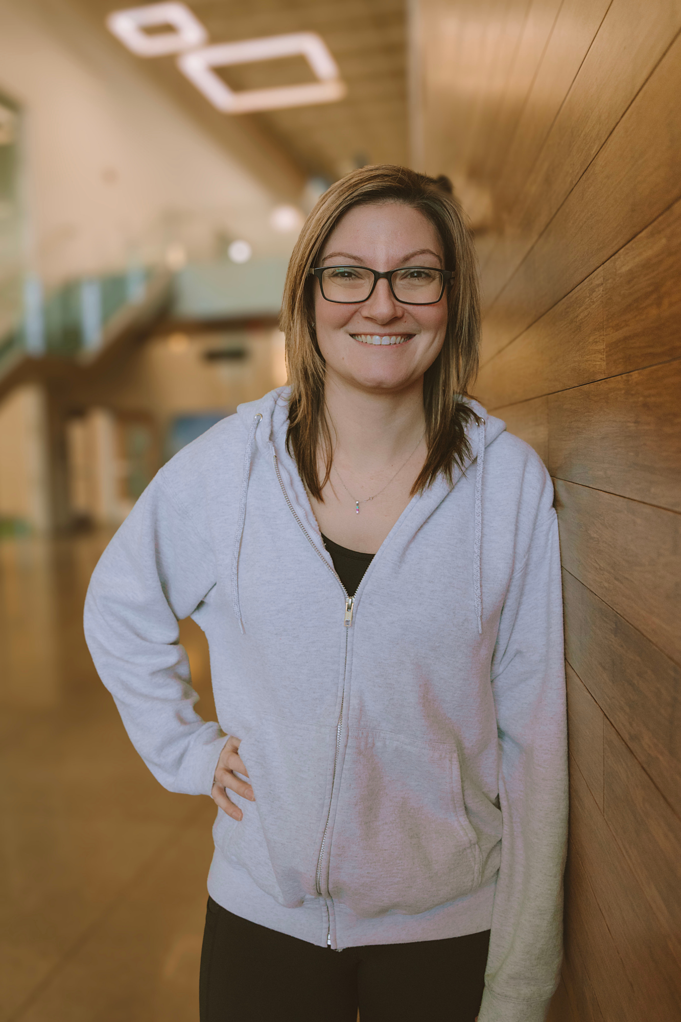 A smiling person wearing glasses and a gray hoodie stands against a wooden wall in a well-lit space.