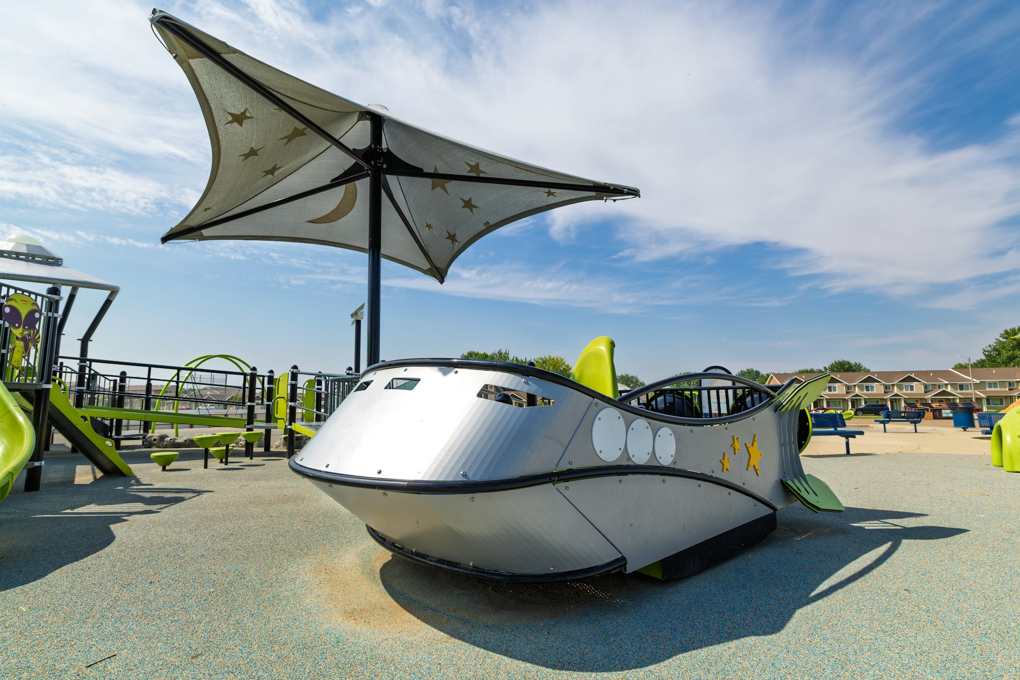 A playground features a boat-shaped structure with a shading canopy, surrounded by slides and other play equipment under a clear sky.