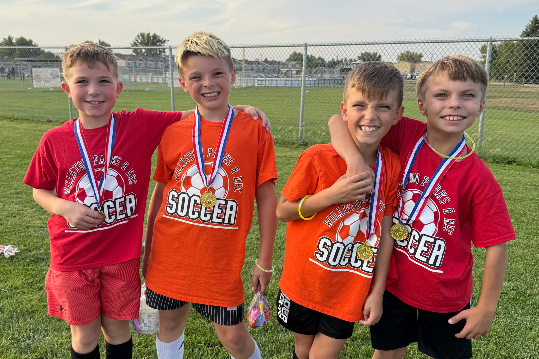 Four kids in soccer shirts with medals, smiling on a grassy field.
