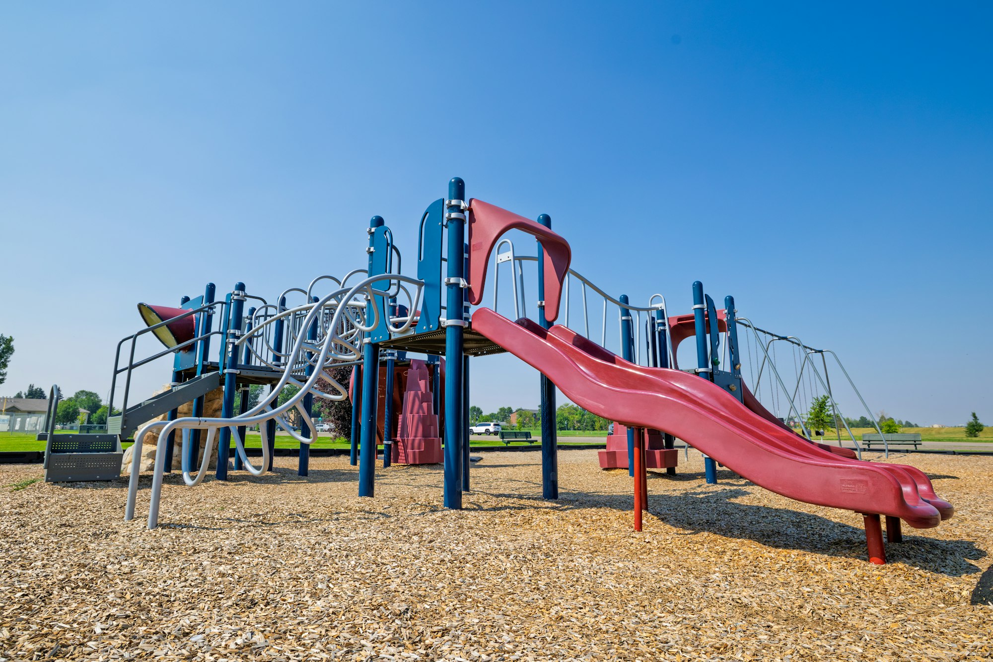 A playground featuring a red slide and climbing structures, set on a wood chip surface under a clear blue sky.