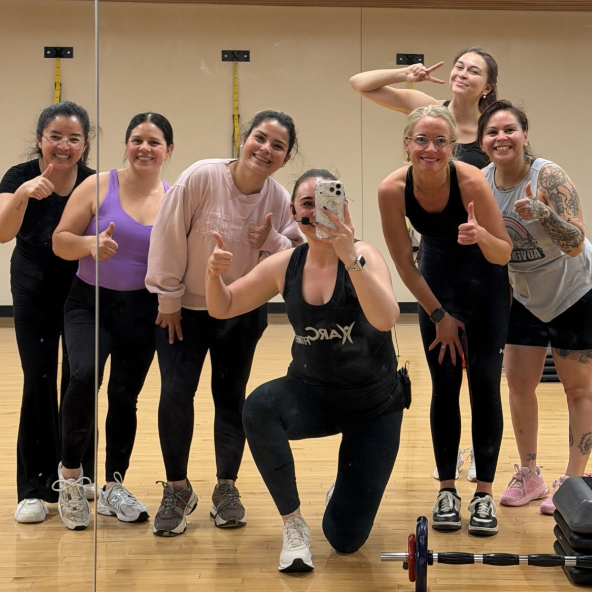 A group of seven women posing happily for a selfie in a fitness studio, showing thumbs up and smiling.
