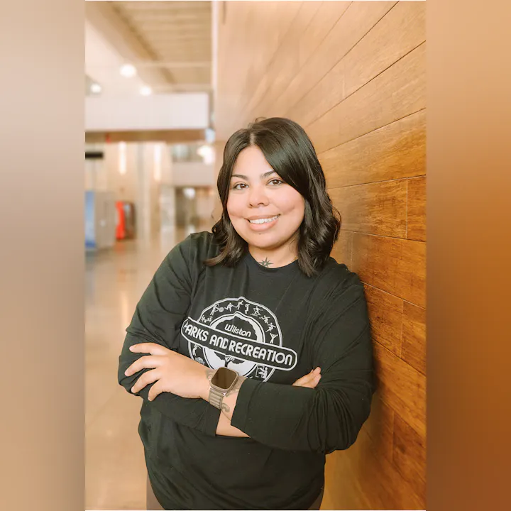 A person smiles while leaning against a wooden wall, wearing a "Wilston Parks and Recreation" shirt in a well-lit indoor space.