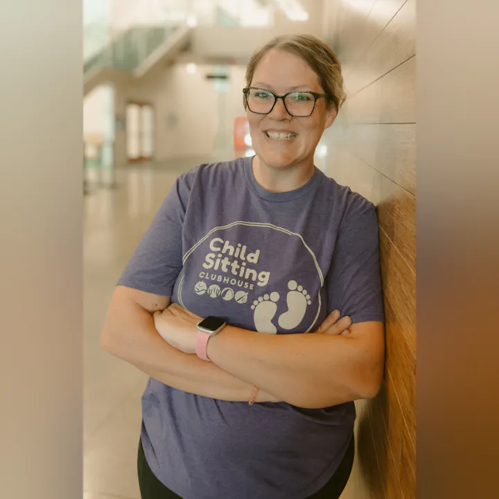A smiling woman in a purple "Child Sitting Clubhouse" shirt stands with arms crossed against a wooden wall indoors.