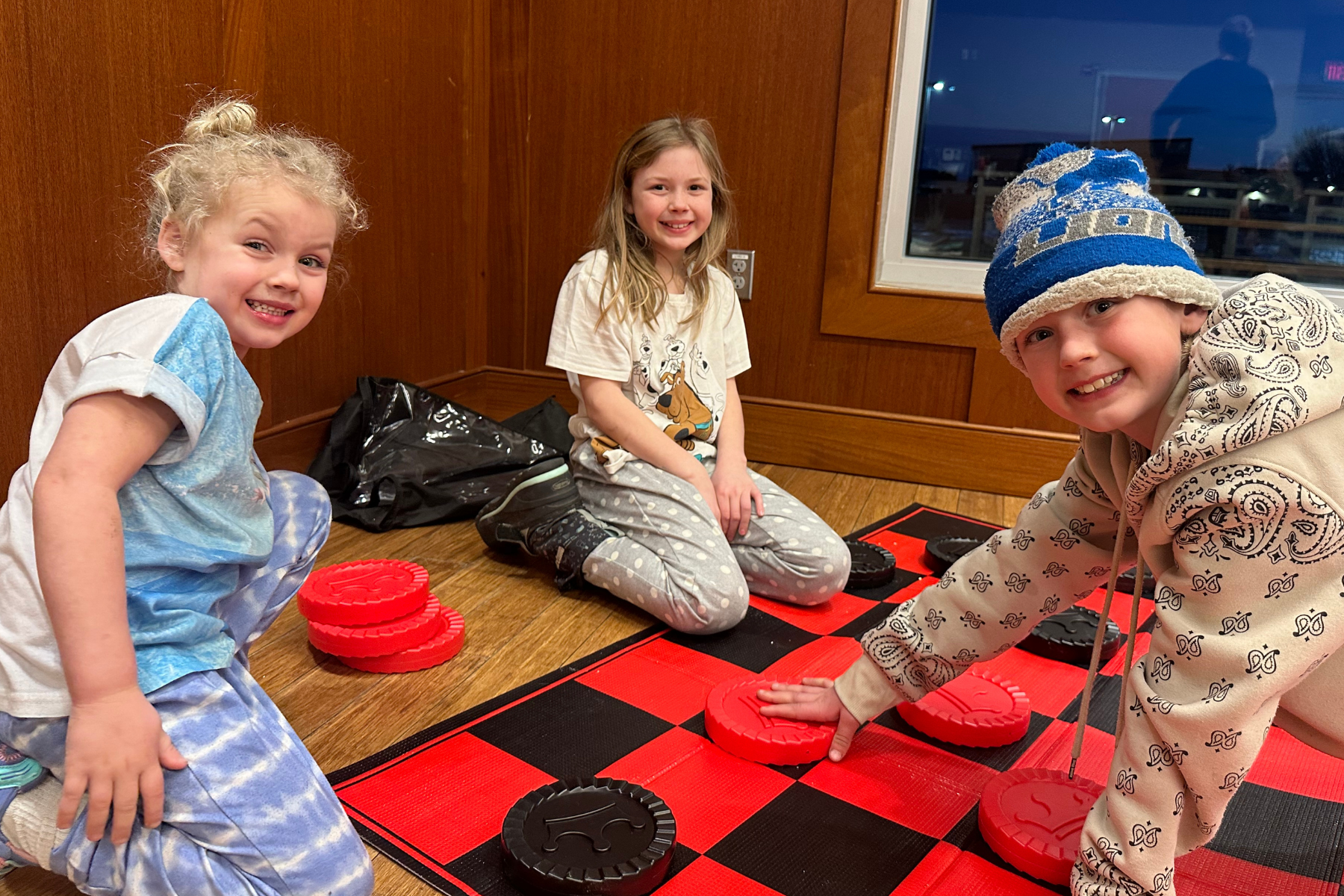 Three children are playing a giant checkers game on a red and black board, smiling and enjoying their time together.