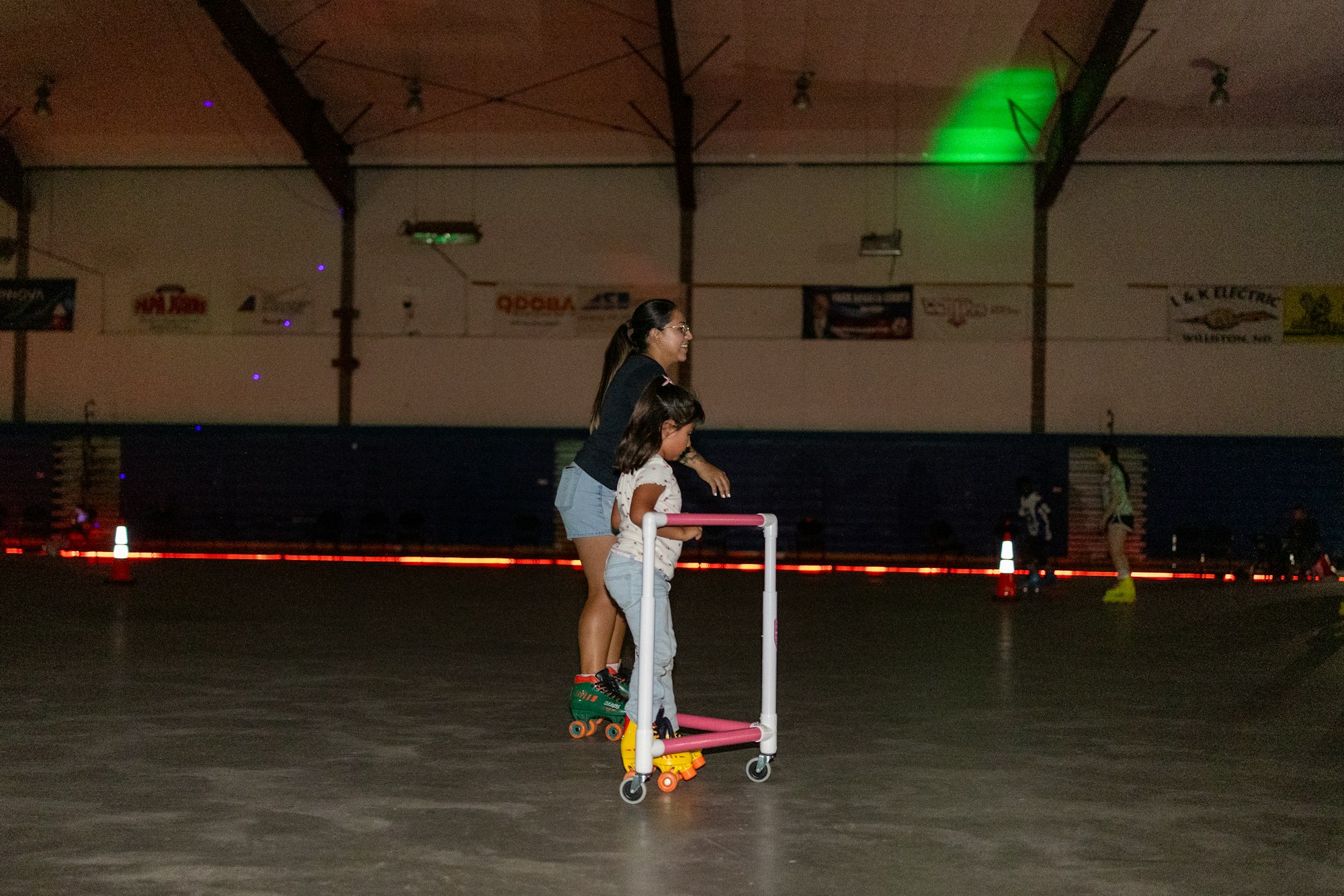 Two people roller skating, one using a support frame, in an indoor rink with colorful lights.