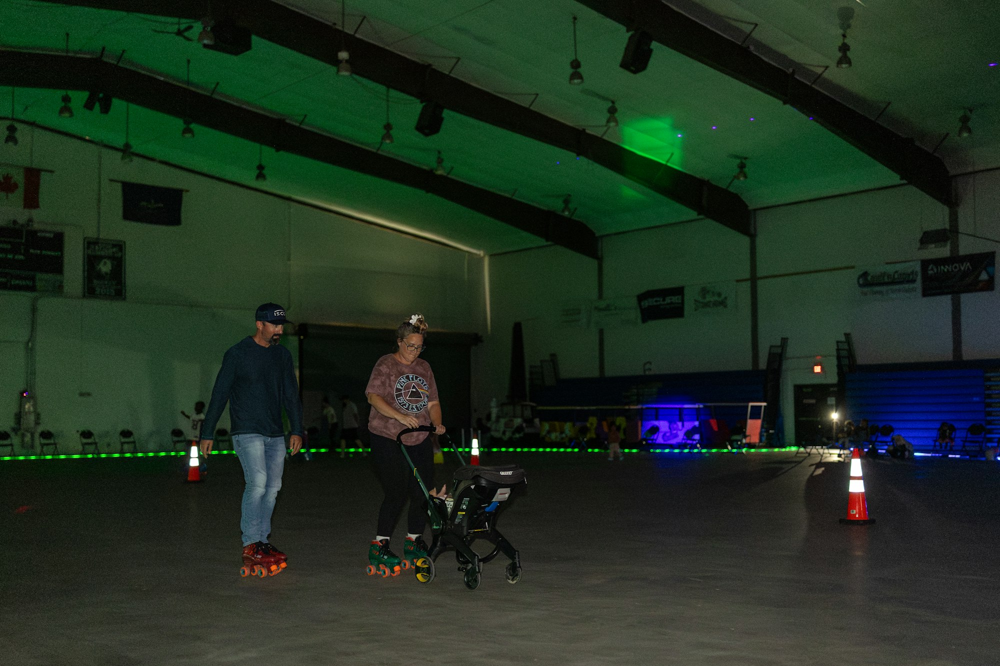 Two people in roller skates inside a dimly lit rink, one pushing a stroller.