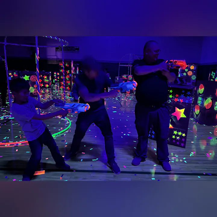 Three people are holding toy blasters in a neon, glow-in-the-dark environment with colorful decorations and confetti on the floor.