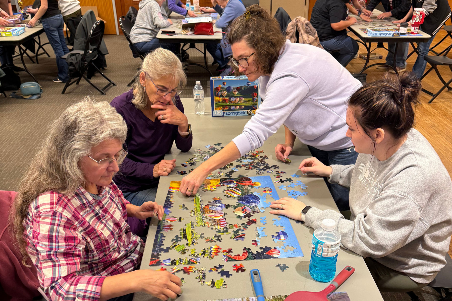 A group of four women work together on a colorful jigsaw puzzle in a lively setting with other puzzles nearby.