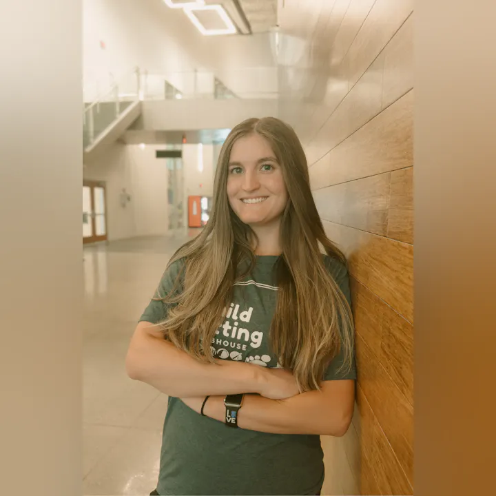 A woman with long hair stands smiling against a wooden wall in a bright indoor space, wearing a casual t-shirt.