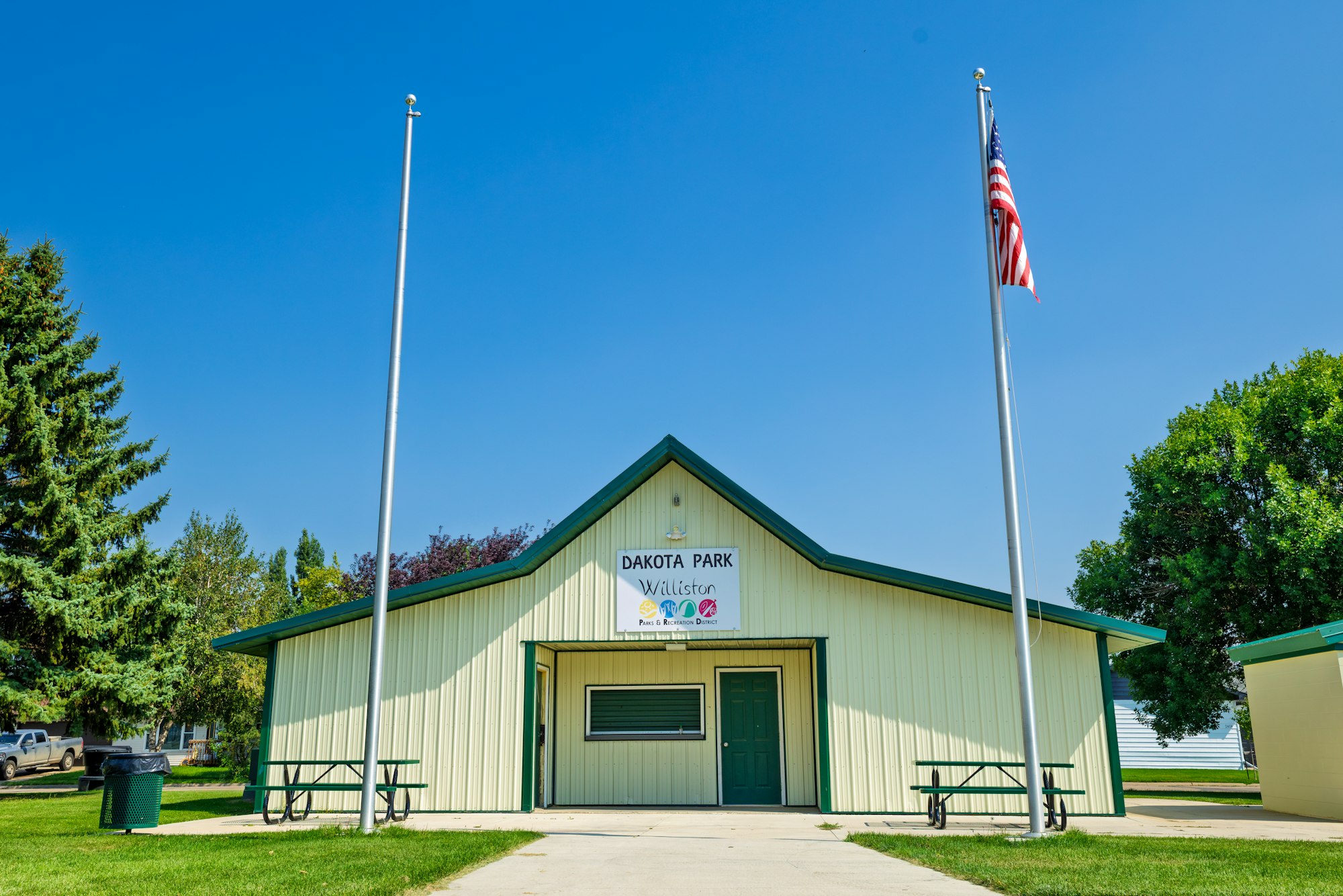 The image shows Dakota Park in Williston, featuring a building, flagpole with an American flag, and picnic tables.