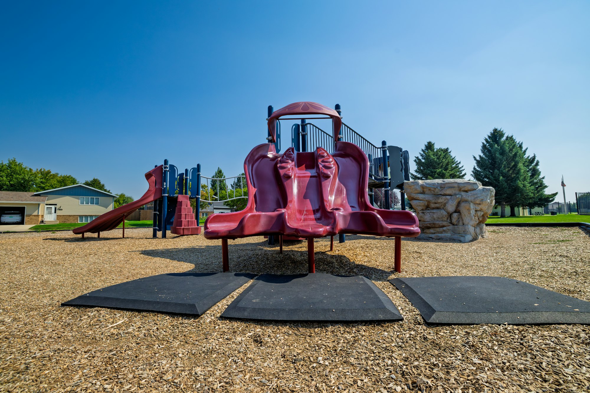 A playground featuring two red slides, climbing stairs, and a rock formation, set against a clear blue sky.