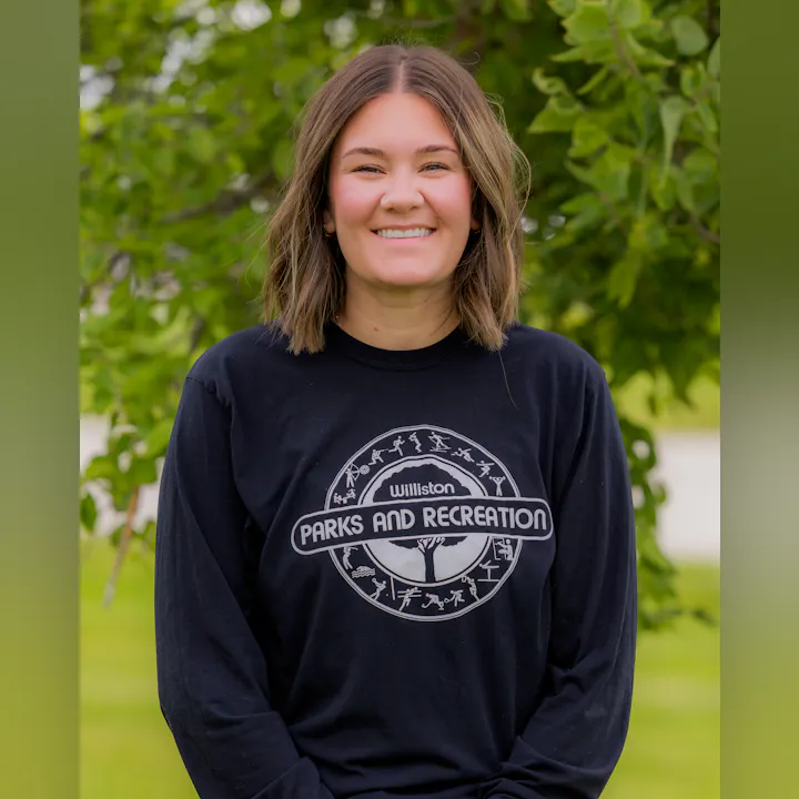 A smiling person wearing a black long-sleeve shirt with "Williston Parks and Recreation" printed on it, against a green backdrop.