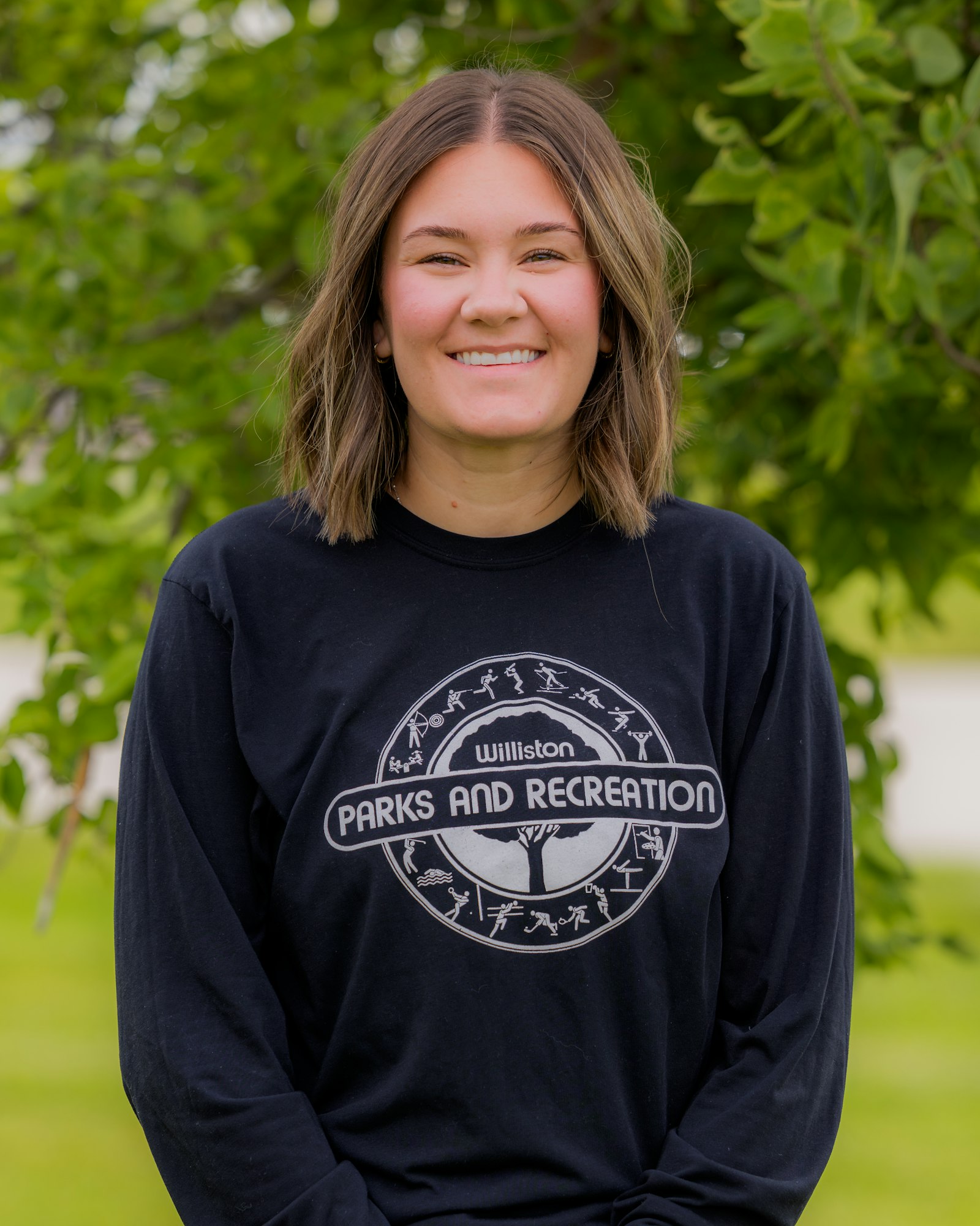 A smiling person wearing a black long-sleeve shirt with "Williston Parks and Recreation" printed on it, against a green backdrop.