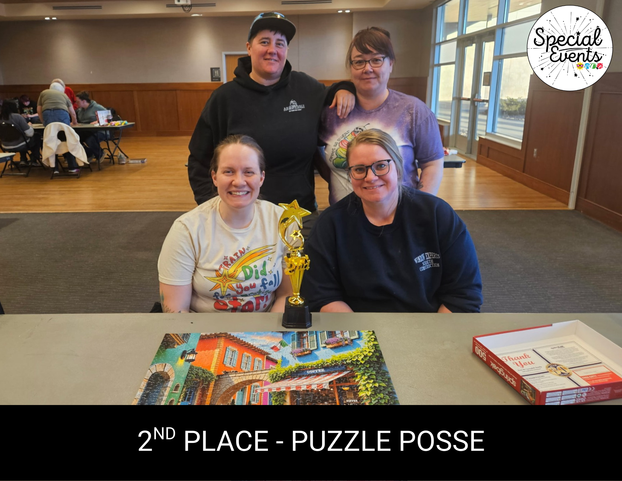 A group of four women poses with a trophy for 2nd place at a puzzle event, with a completed puzzle on the table.