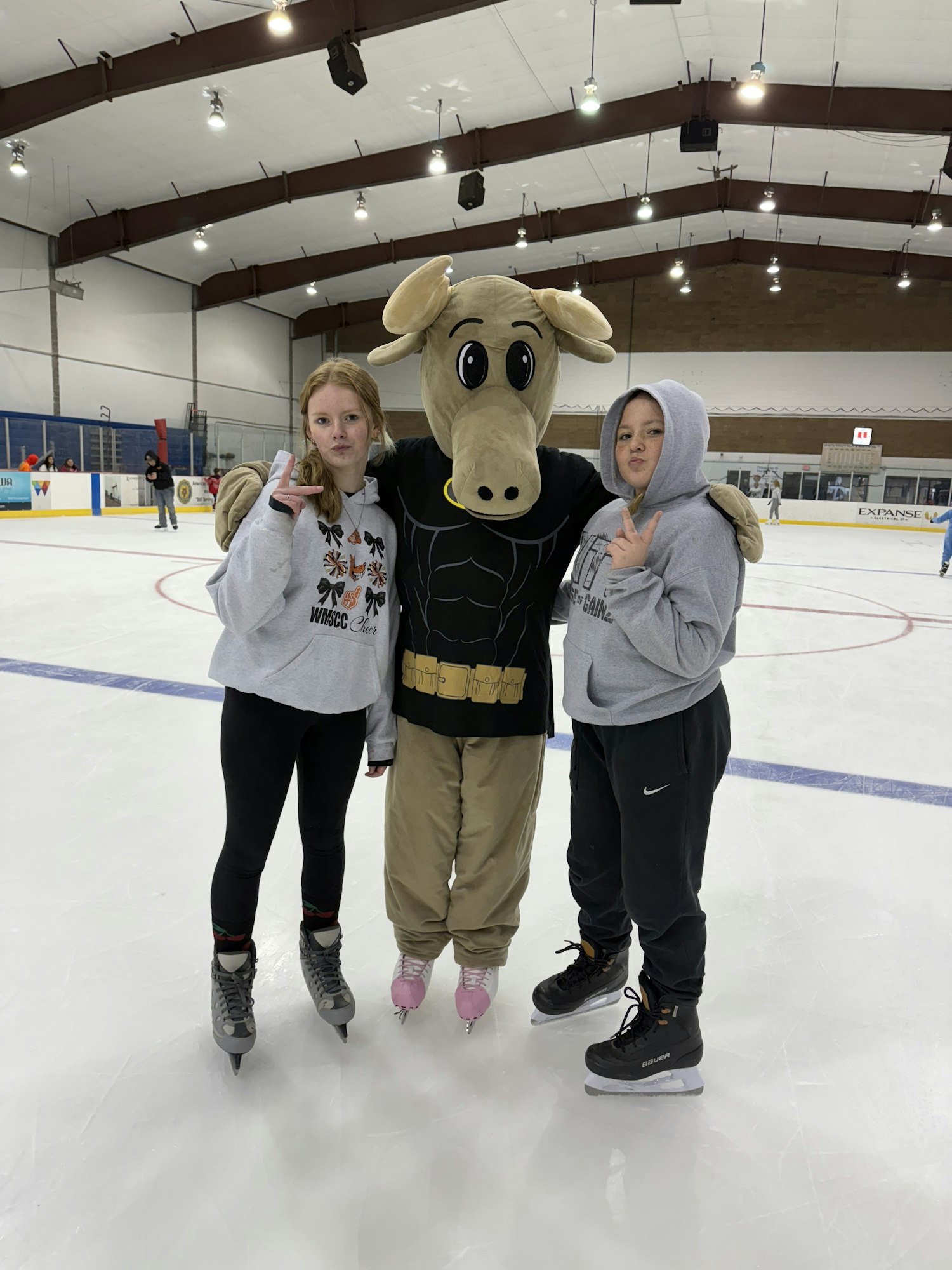 Two girls and a mascot in a moose costume pose together on an ice rink. All are dressed casually, smiling.